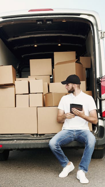 A man wearing a black cap, white t-shirt, and blue jeans is seated on the pavement beside the open rear of a van during a house removal. The van’s interior is filled with stacked cardboard boxes of various sizes, some of which are sealed with packing tape, indicating packing and moving preparations. The boxes are arranged on the van floor, with larger boxes positioned at the back and smaller ones towards the front, suggesting an organized loading process. The man is holding a tablet or clipboard, possibly checking inventory or coordinating the relocation process, as part of furniture transport and packing activities. The environment appears to be an outdoor area near a property, with natural daylight illuminating the scene, reflecting the typical loading phase in a home relocation carried out by Man with Van Knightsbridge, a professional removals service.