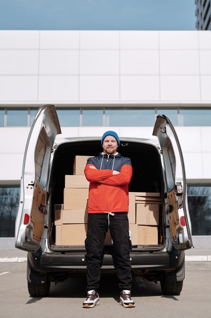 A man wearing a blue beanie, red and navy jacket, and black trousers stands with arms crossed in front of an open van loaded with several cardboard boxes, which are arranged neatly inside the vehicle. The van is parked outside a modern building with a white tiled facade, large glass windows, and horizontal metal panels. The scene is set during daylight, with clear weather and natural lighting. Visible on the pavement are the rear doors of the van and a small portion of the surrounding urban environment. This image illustrates the packing and loading process involved in home relocation services provided by Man with Van Knightsbridge, highlighting the transportation of household items as part of a professional removals operation.