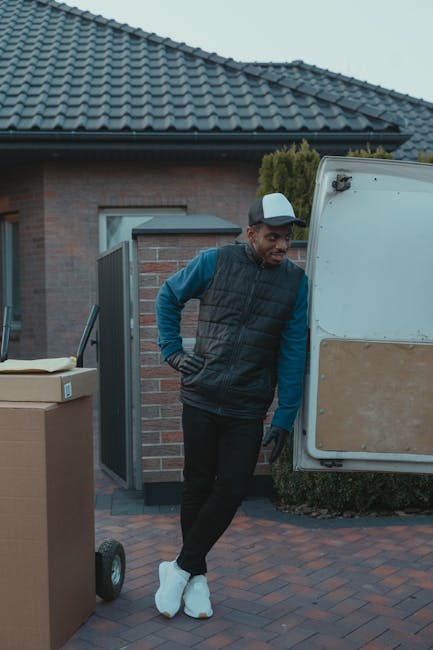 A man wearing a black cap, white t-shirt, and blue jeans is seated on the pavement beside the open rear of a van during a house removal. The van’s interior is filled with stacked cardboard boxes of various sizes, some of which are sealed with packing tape, indicating packing and moving preparations. The boxes are arranged on the van floor, with larger boxes positioned at the back and smaller ones towards the front, suggesting an organized loading process. The man is holding a tablet or clipboard, possibly checking inventory or coordinating the relocation process, as part of furniture transport and packing activities. The environment appears to be an outdoor area near a property, with natural daylight illuminating the scene, reflecting the typical loading phase in a home relocation carried out by Man with Van Knightsbridge, a professional removals service.