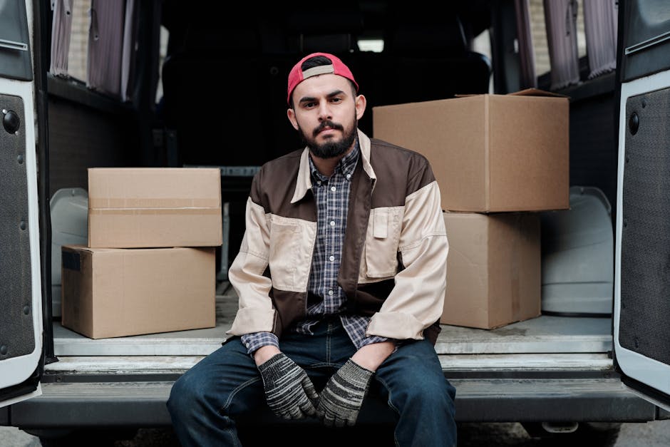 A young man with a beard, wearing a red cap, a checkered shirt, and a multi-toned beige and brown jacket, is sitting on the rear bumper of a moving van with its back doors open. Inside the van, there are several cardboard boxes of different sizes, some stacked on top of each other, and visible wrapping materials such as plastic and fabric padding. The man is wearing striped gloves, with his hands resting on his knees, and is looking directly at the camera with a neutral expression. The vehicle is parked on a street, and the scene captures the loading process involved in home relocation or furniture transport, with the focus on the organized packing and the vehicle ready for moving services as provided by Man with Van Knightsbridge.