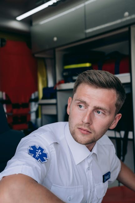 A male paramedic with short brown hair and light skin, wearing a white uniform with a blue medical emblem on his sleeve, is inside an ambulance or medical vehicle. The background shows shelves with red and yellow medical equipment, plastic storage containers, and other emergency supplies. The man appears to be in a state of alertness or contemplation, possibly during a house removal or emergency relocation process involving Man with Van Knightsbridge, as suggested by the context of moving and transportation services. The interior lighting is bright, highlighting the details of his facial expression and the equipment around him, reflecting the professional environment associated with rapid response and logistics support for home relocations or urgent removals.