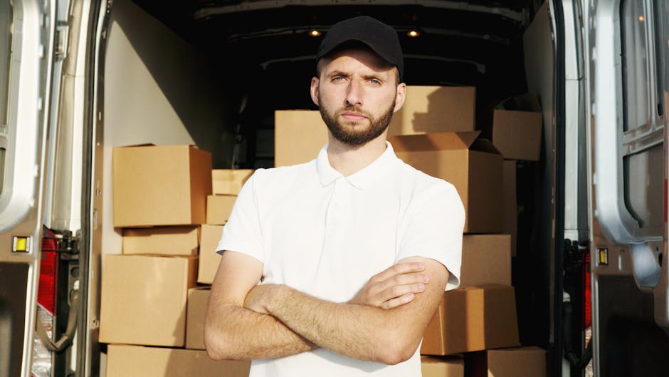A young man with a beard, wearing a black cap and white polo shirt, stands with arms crossed in front of an open van loaded with neatly stacked cardboard boxes. The boxes vary in size and are secured with plastic wrap. The van, parked on a street, has its rear doors open, revealing the interior filled with additional packaging materials. The scene captures the loading process during a typical house removal or home relocation, with the man representing a professional mover from Man with Van Knightsbridge, involved in furniture transport and packing services, adhering to Knightsbridge council permit rules for removals in RBKC.