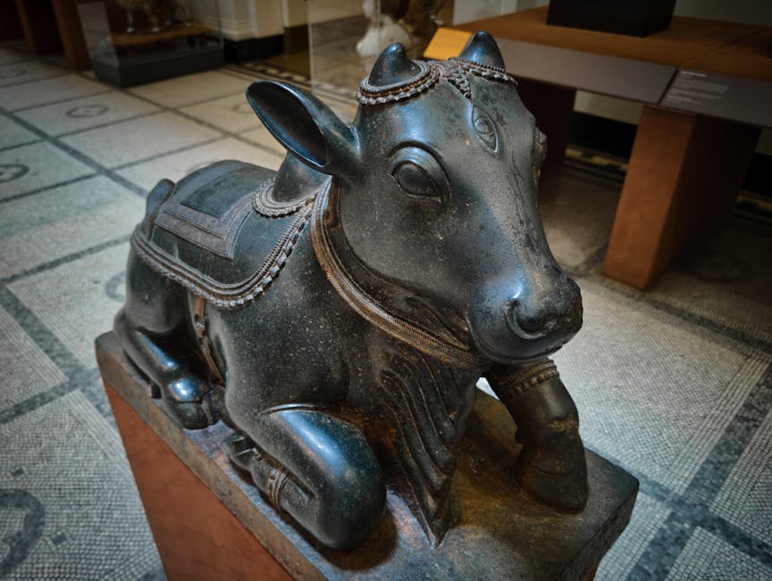 A detailed view of a dark metal Yaksha-style sculpture of a crouching cow, situated indoors on a wooden pedestal. The sculpture features intricate decorative elements, including a saddle with ornate patterns and a collar around its neck, all crafted with fine detailing. The sculpture has a smooth, polished surface with visible signs of patina and aging, giving it an antique appearance. The background includes a patterned carpet, part of a room setting, with a wooden table partially visible, indicating a curated indoor environment. The scene captures the craftsmanship of the sculpture, emphasizing its cultural and artistic significance, which may be of interest in the context of house or furniture relocation, packaging, and transportation for art objects, as handled by companies like Man with Van Knightsbridge.