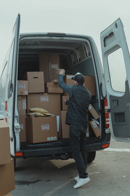 A person wearing a dark jacket, light-colored sneakers, and a cap is arranging and carrying cardboard boxes inside the open cargo area of a white van, which is parked on the pavement near a building. The van's interior is filled with stacked boxes of various sizes, some wrapped in protective packaging materials. The scene depicts a loading process during a home relocation or furniture transport task, with the individual preparing for the transportation of household items. The background shows an outdoor urban environment with a light cloudy sky, indicating daylight hours. Man with Van Knightsbridge, a professional removals company, is visible in the context of moving services and packing and loading procedures.