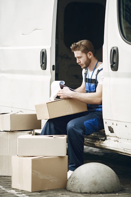 A professional mover from Man with Van Knightsbridge is loading numerous cardboard boxes into the back of a large, open delivery van parked on a city street, possibly during a house removal or relocation process. The van's interior is filled with stacked boxes, some positioned on a trolley that the mover is using to transport items inside. The mover, dressed in dark clothing with a jacket, is seen from behind as he arranges the boxes within the vehicle, which is situated on a paved pavement near trees and modern buildings. An additional cardboard box is on the ground beside the van, ready to be loaded or unloaded. The environment suggests an urban setting, with the van securely positioned for efficient furniture transport, packing, and moving activities. The scene captures a typical loading process involved in home relocation services provided by Man with Van Knightsbridge, illustrating the careful handling of items during furniture transport and the preparation for further moving logistics.