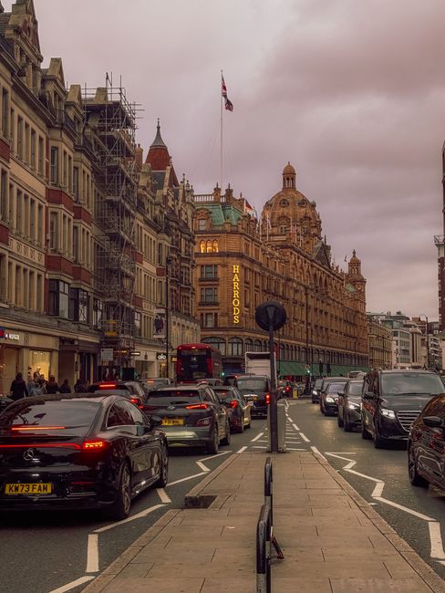 A busy street scene in Knightsbridge, London, showing a line of parked cars and moving vehicles, including a black car and a red double-decker bus. The buildings lining the street are historic, with o
