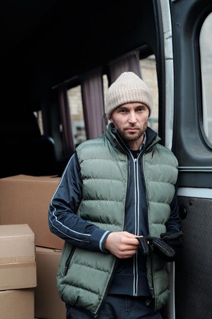 A man with a serious expression, wearing a light grey knit beanie and a padded green and navy blue jacket, stands inside the open sliding door of a moving van, holding a black strap. Behind him, the interior of the van is visible with several packed cardboard boxes stacked along the side, prepared for a domestic home relocation. The van appears to be parked on a residential street, indicated by blurred buildings and street elements visible through the windows. The scene captures the loading or unloading process as part of a professional furniture transport or packing and moving service, with focus on secure item handling during a house removal. The presence of multiple boxes and the professional environment subtly highlights the logistical aspect of local or long-distance removals handled by [COMPANY_NAME], supporting effective house moving services near Sloane Street and Knightsbridge.