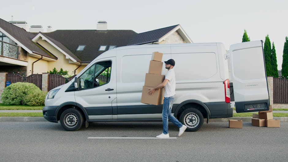 A man wearing casual clothing and a cap is standing outside a white medium-sized van parked along a residential street, carrying three cardboard boxes stacked vertically. Additional cardboard boxes are placed on the pavement beside the van, some stacked and some lying flat. The van is in the process of loading or unloading, as part of a home relocation or furniture transport operation carried out by Man with Van Knightsbridge, visible from their service page near Sloane Street. The background features a modern detached house with a dark tiled roof, large windows, and a garden with neatly trimmed bushes and tall green trees. The environment is well-lit with natural daylight, and the scene captures the typical logistics involved in packing and moving, with the van positioned close to the property for efficient loading or unloading during a domestic removal or moving service.