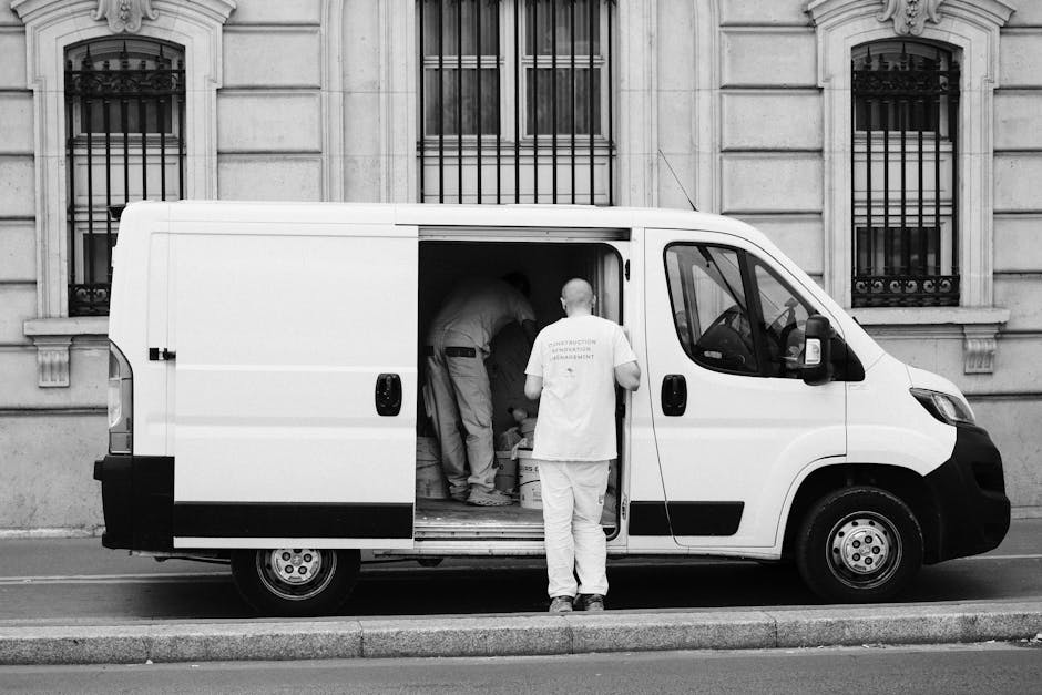 A man with a serious expression, wearing a light grey knit beanie and a padded green and navy blue jacket, stands inside the open sliding door of a moving van, holding a black strap. Behind him, the interior of the van is visible with several packed cardboard boxes stacked along the side, prepared for a domestic home relocation. The van appears to be parked on a residential street, indicated by blurred buildings and street elements visible through the windows. The scene captures the loading or unloading process as part of a professional furniture transport or packing and moving service, with focus on secure item handling during a house removal. The presence of multiple boxes and the professional environment subtly highlights the logistical aspect of local or long-distance removals handled by [COMPANY_NAME], supporting effective house moving services near Sloane Street and Knightsbridge.