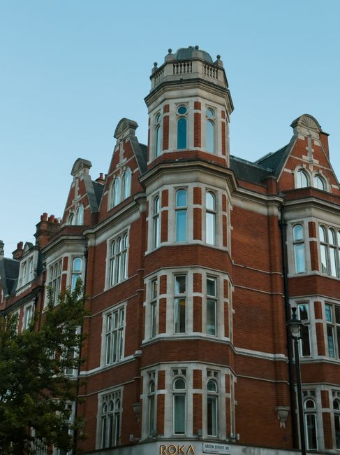 A busy street scene in Knightsbridge, London, featuring a row of historic Victorian-style buildings with ornate facades and large windows, some with rooftop and decorative turrets. Parked cars, includ