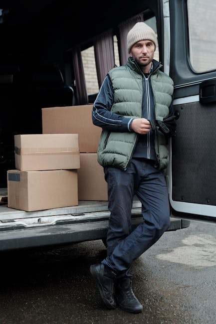 A man wearing a light grey beanie, green puffer vest, and dark trousers is standing at the rear of a van with the sliding door open. Inside the van, several cardboard boxes of varying sizes are visible, some with black tape or labels. The man is holding a small black object, possibly a remote or a phone, and appears to be taking a break during a home relocation process. The van is parked on a street, and the background shows an overcast sky, suggesting a typical day for furniture transport or packing and moving services. The scene reflects the loading process involved in professional removals, with the focus on the secure packing of household items for transportation, supported by the presence of packaging materials such as cardboard boxes and the vehicle used by [COMPANY_NAME] for residential removals.
