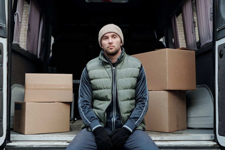 A young man wearing a beige knit beanie, a green padded vest over a navy blue tracksuit top, and black gloves is seated inside the open back of a black van, which is loaded with several cardboard boxes of various sizes. The boxes are taped shut and some are stacked, positioned on a metal flooring surface within the vehicle. The van is parked on a street, with a building or house visible in the background. The interior of the van shows a loading area where furniture and packing materials are organized for a home relocation or furniture transport service, with the vehicle used for professional removals such as those provided by Man with Van Knightsbridge.