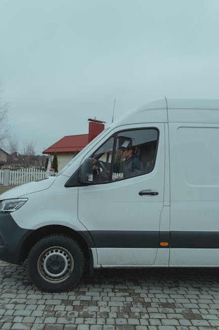 A man wearing a light grey beanie, green puffer vest, and dark trousers is standing at the rear of a van with the sliding door open. Inside the van, several cardboard boxes of varying sizes are visible, some with black tape or labels. The man is holding a small black object, possibly a remote or a phone, and appears to be taking a break during a home relocation process. The van is parked on a street, and the background shows an overcast sky, suggesting a typical day for furniture transport or packing and moving services. The scene reflects the loading process involved in professional removals, with the focus on the secure packing of household items for transportation, supported by the presence of packaging materials such as cardboard boxes and the vehicle used by [COMPANY_NAME] for residential removals.