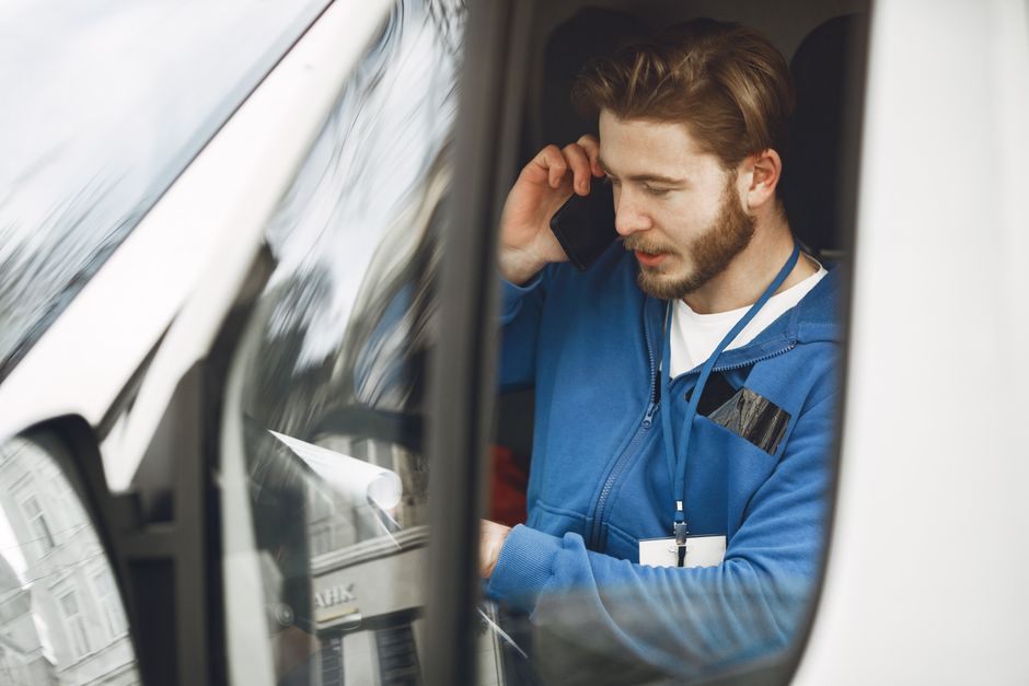A man with a beard and brown hair, wearing a blue zip-up jacket and a lanyard with an ID badge, is sitting inside a white van, likely during a house or office relocation. He is holding a mobile phone to his ear with his right hand and appears to be engaged in a conversation. Inside the van, various packing materials are visible, including cardboard boxes, plastic wrapping, and furniture blankets used for protection during furniture transport. The interior of the van shows part of the dashboard and a window through which reflections of nearby buildings and the urban environment can be seen. The man is positioned near the doorway of the van, which is open, revealing the moving process. This scene aligns with professional removal services like those offered by Man with Van Knightsbridge, supporting packing, loading, and transportation of household or office items in a detailed and factual manner, emphasizing efficient and careful moving logistics.