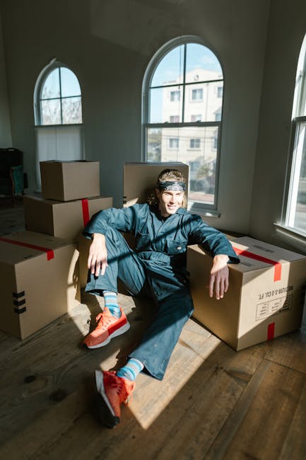 A young man dressed in casual work attire, including a dark blue jumpsuit, sits on a wooden floor surrounded by multiple large cardboard moving boxes secured with red packing tape inside a bright room with tall, arched windows. The windows allow natural daylight to illuminate the scene, revealing an exterior view of nearby buildings. The man is smiling and relaxed, with one arm resting on a box and the other on his knee. In the background, there are additional boxes placed on the floor, suggesting an ongoing home relocation or packing process. The setting indicates a furniture transport and packing stage for a house move, with visible packaging materials and a spacious, well-lit environment. Man with Van Knightsbridge provides professional removals services, including furniture transport and packing, essential for a smooth house relocation process.
